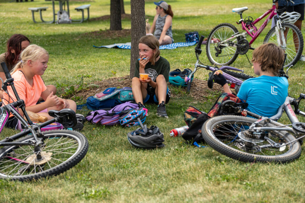 Children sitting down next to a tree surrounded by bikes, one of them holding a fruit cup.