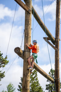 A girl with a light blue helmet, climbing up a wooden structure.