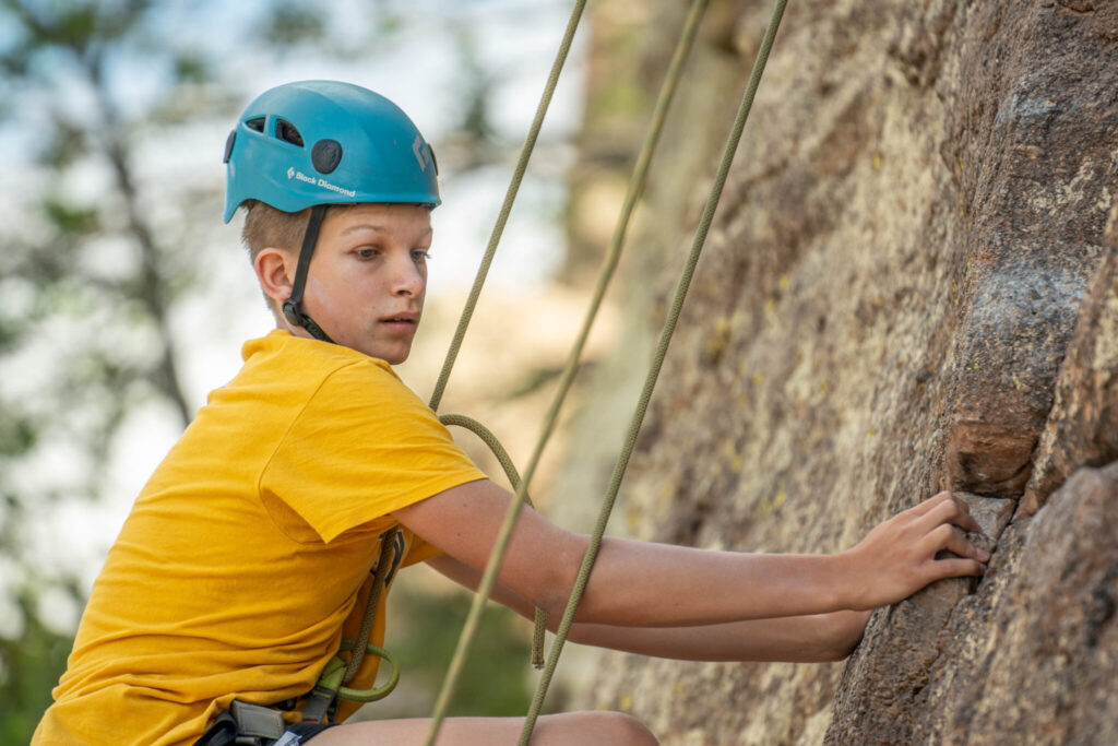 A Child with a blue helmet looking at a crack in the boulder while climbing.