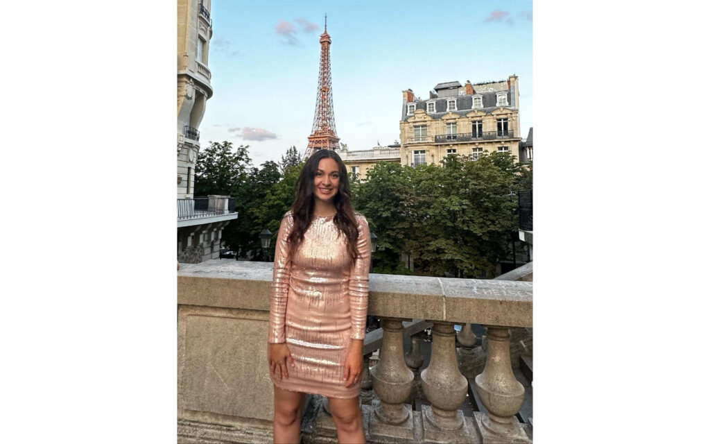 Natalie, in a sequined dress, standing in front a set of buildings and the Eiffel tower.