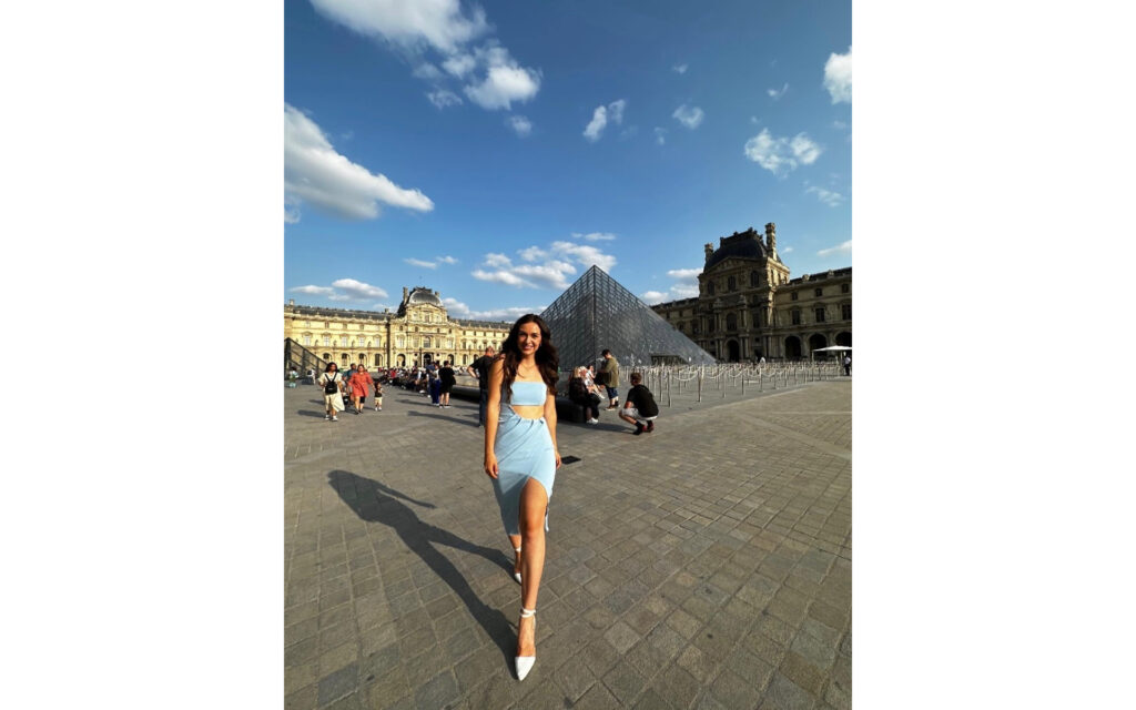 Natalie, in a blue dress, standing in the Louvre courtyard, in front of the pyramid.