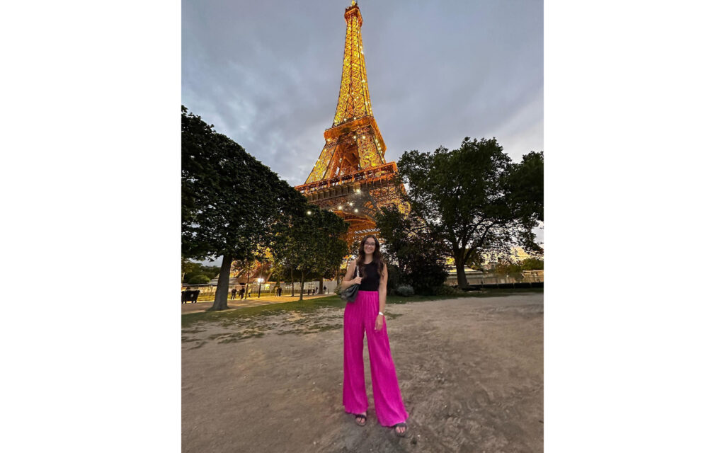Natalie, in pink pants, standing in front of the Eiffel Tower, with it's lights on.