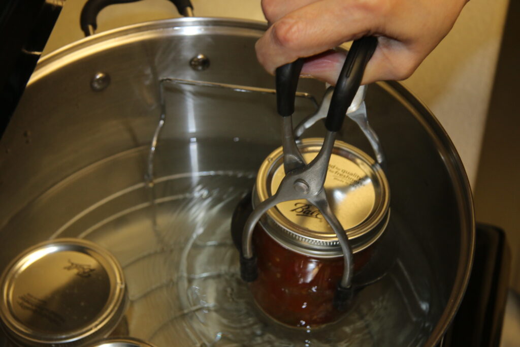 A cooker holding hot water with a hand removing a jar with a tool