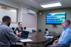 Three people sitting around a table with microphones and a TV in the background with a graphic reading 'Health and Human Sciences Matters'