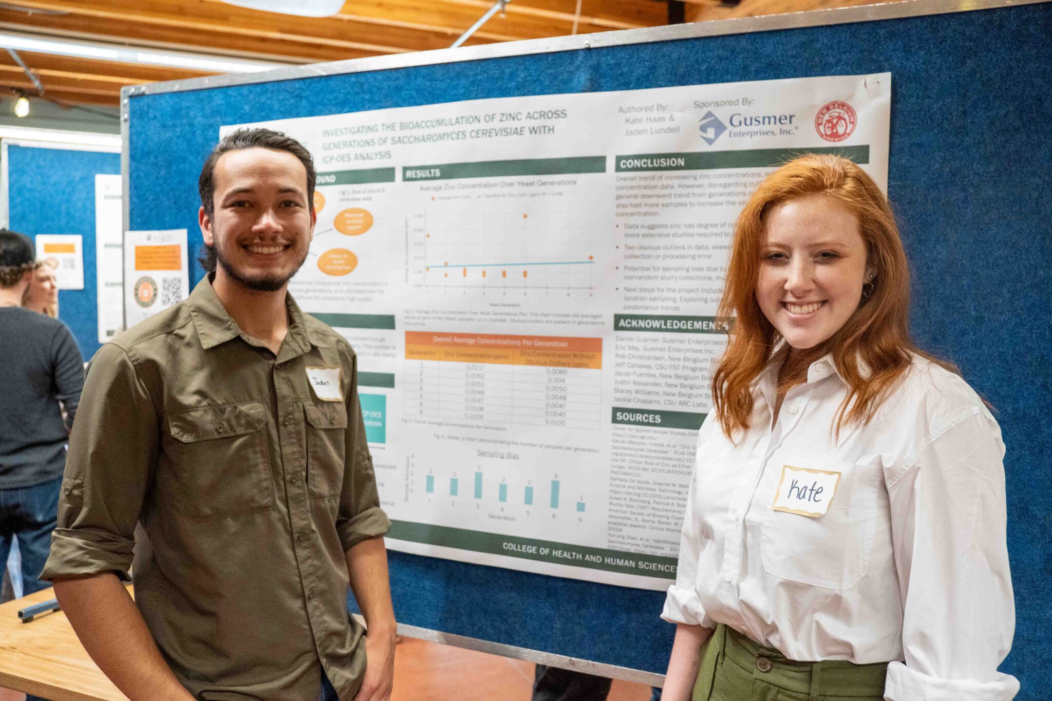 A red-haired woman and a man smile in front of their poster presentation at the FST senior capstone show