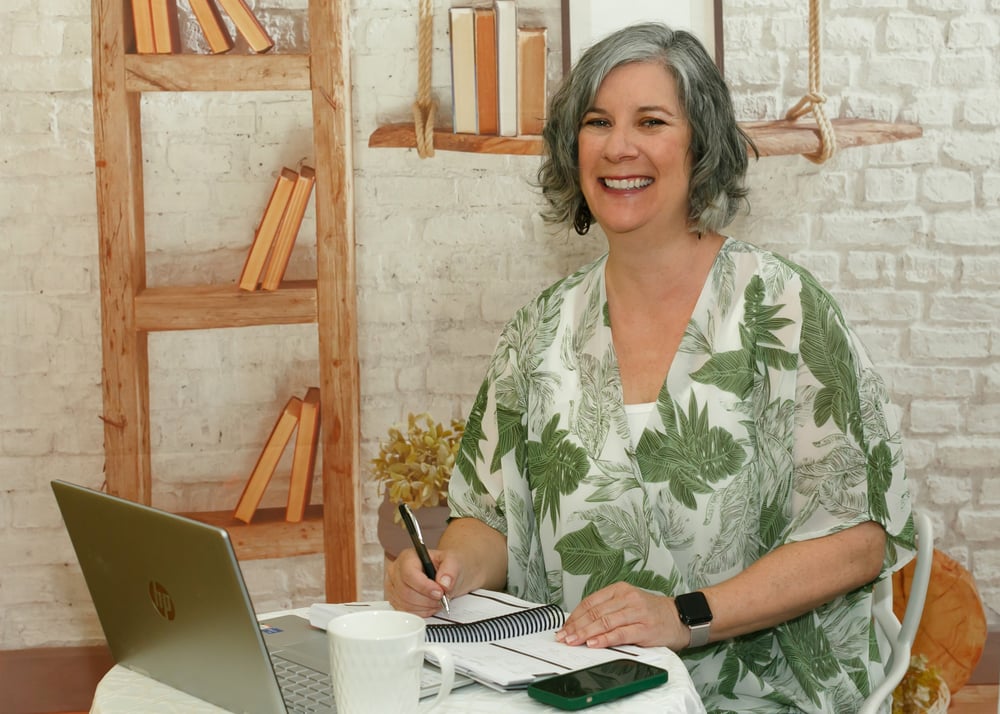 Suzie Scher sitting at a small table, smiling, with a laptop and notebooks in front of her and a white brick wall in the background