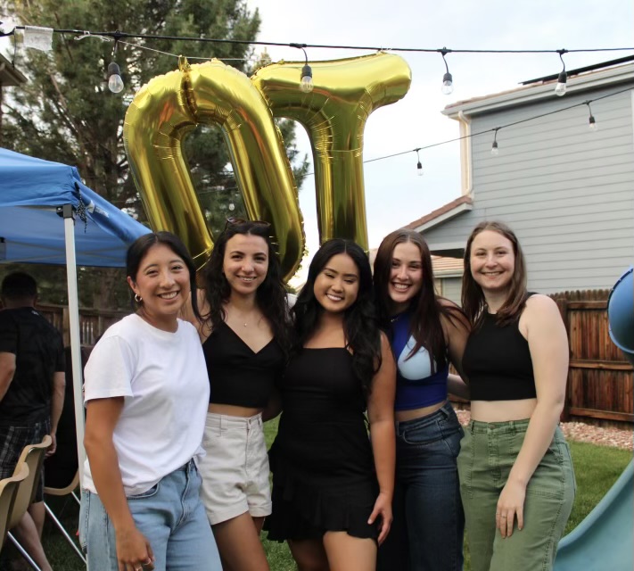 Five women pose for a photo in a backyard with letter balloons "OT" behind them.
