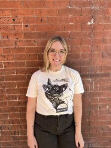 Lauren poses in front of a brick wall. She is wearing glasses and a white graphic t-shirt.