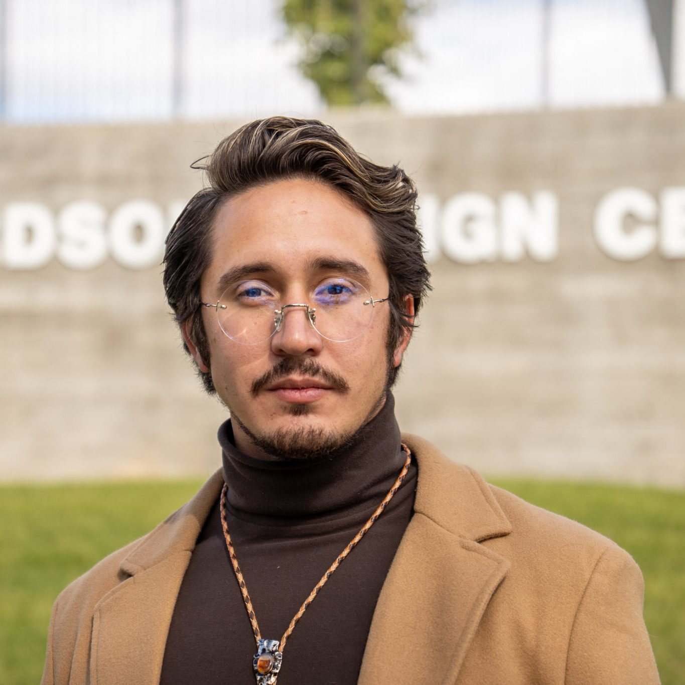Payton wears a brown turtleneck and light brown jacket with a necklace and glasses outside the Nancy Richardson Design Center.