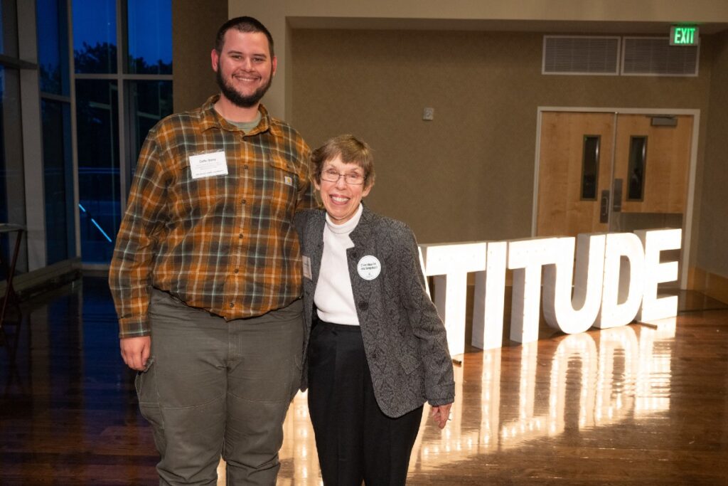 A tall man and an older woman stand in front of a lighted "gratitude sign"