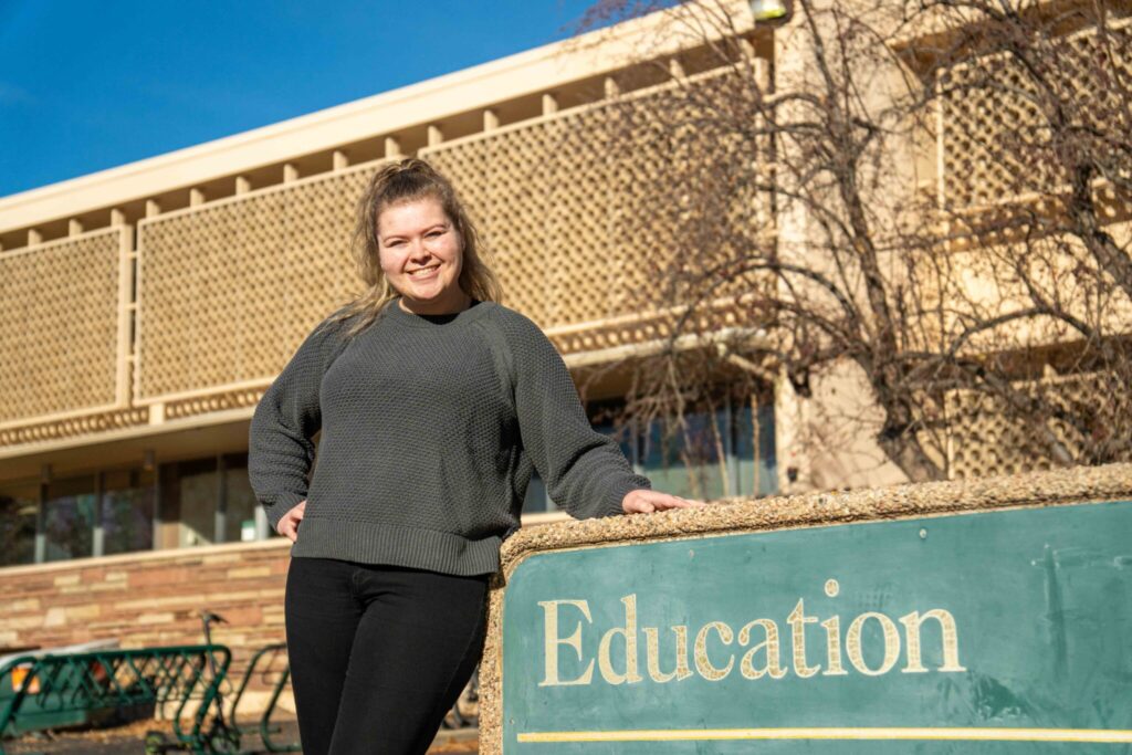 Eden wears a gray sweater and black leggings and boots. She is posing outside the Education building, leaning on the sign.