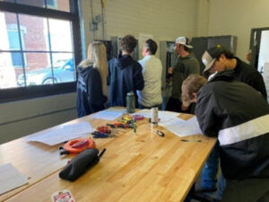 Five students and 1 instructor looking at an electrical panel on wall