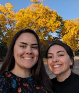Ashley and a female friend pose with yellow fall trees behind them.