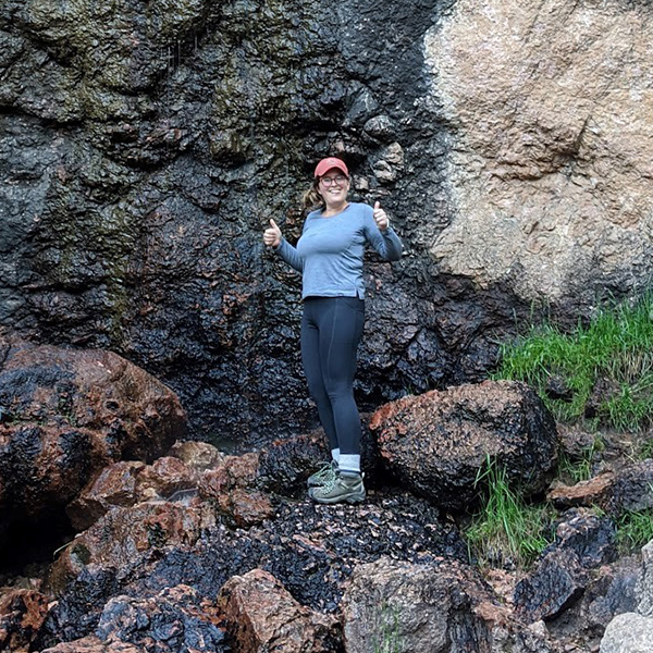Hannah Burke holds up a thumbs up with standing on rocks. She is wearing hiking clothes and a red baseball cap.