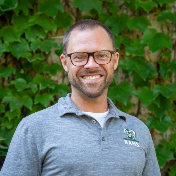 Charlie Hoxmeier smiles in front a wall of greenery wearing a gray CSU Rams polo shirt. He is wearing black glasses.