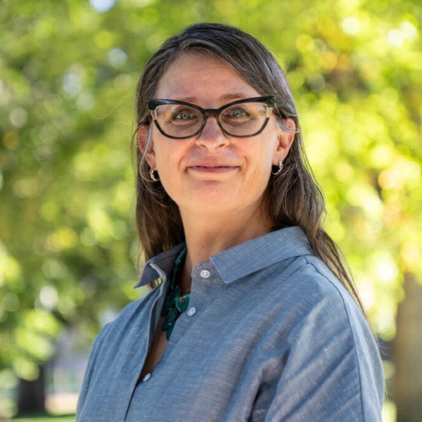Christine Murphy smiles wearing a blue button up shirt and cat-eyed shaped glasses on the Oval.
