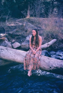 Gail Strid, in a dress and barefoot, seated on a log that crosses a river in Colorado, 1975