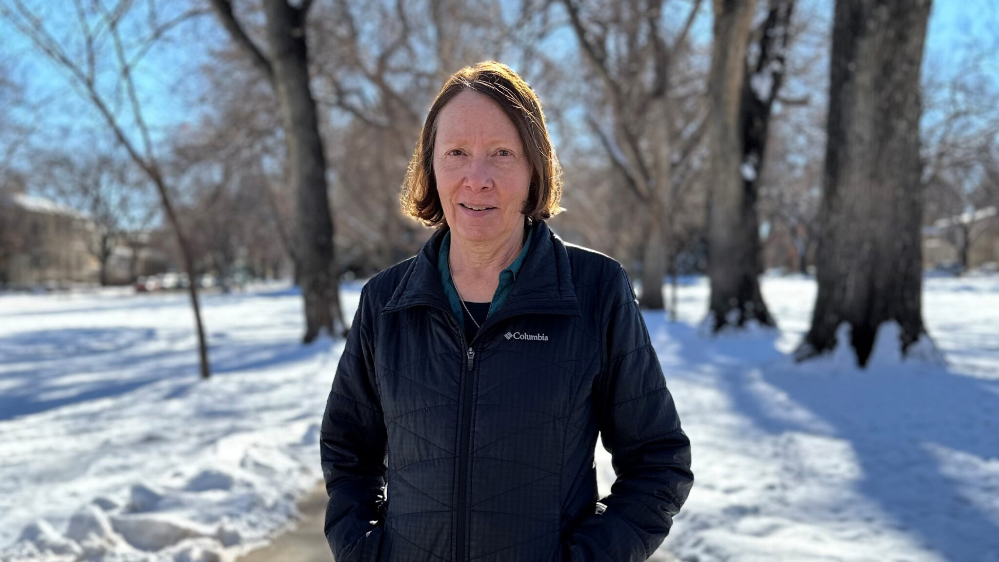 Gail Strid standing on a sidewalk surrounded by snow with leafless elm trees in the background and blue skies
