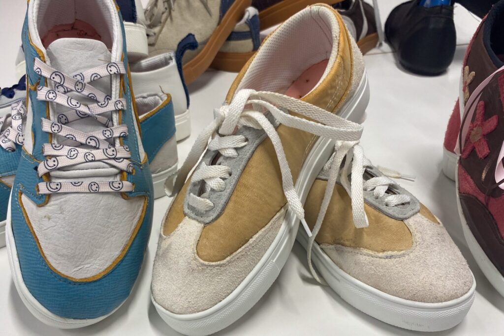 an image showcasing two pairs of shoes created by the students one with gold and white leather and white laces and one with blue and white leather, yellow accents and smiley face laces