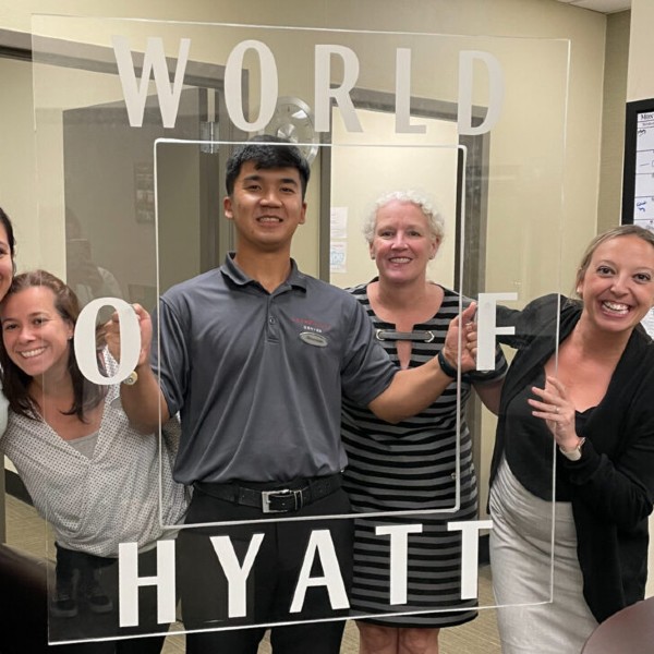 Ilkhomiddin-Nabijonov smiles with 3 female Hyatt staff members holding a plexiglass frame that says " World of Hyatt" around the border.