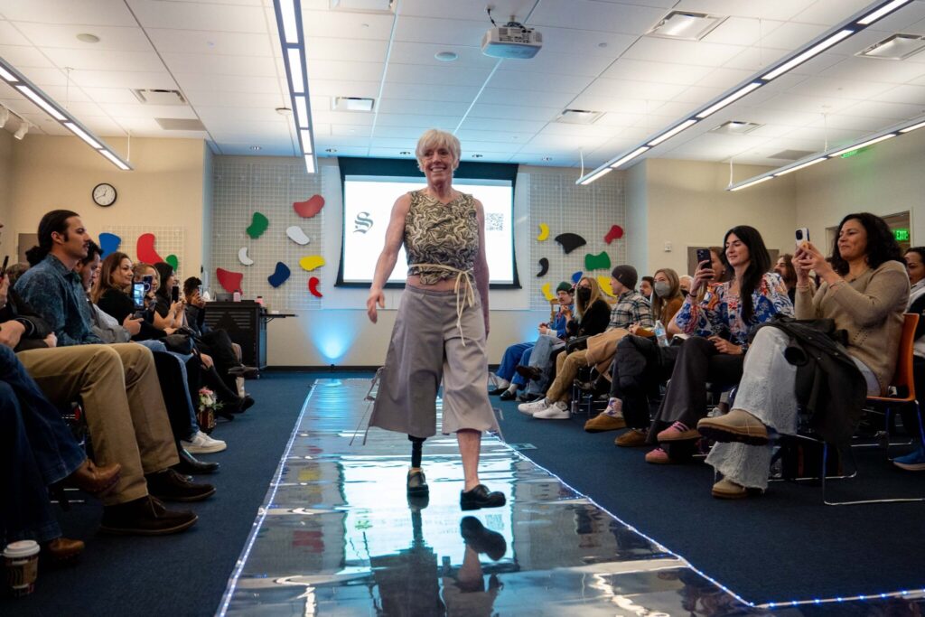 A woman with a prosthetic leg models a pair of capri pants and a tank as she walks down the runway with a large crowd taking photos