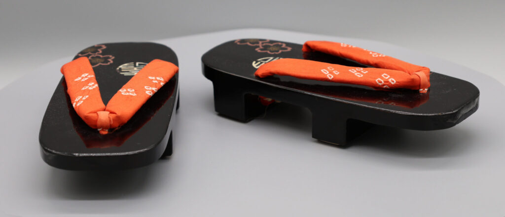 A pair of Japanese Geta sandals sit on a white background. They are black with a orange strap.