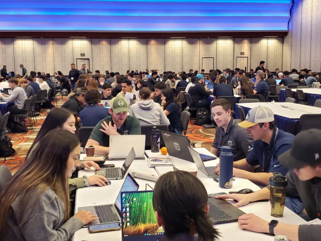 A large convention-style room filled with students sitting at tables working on computers