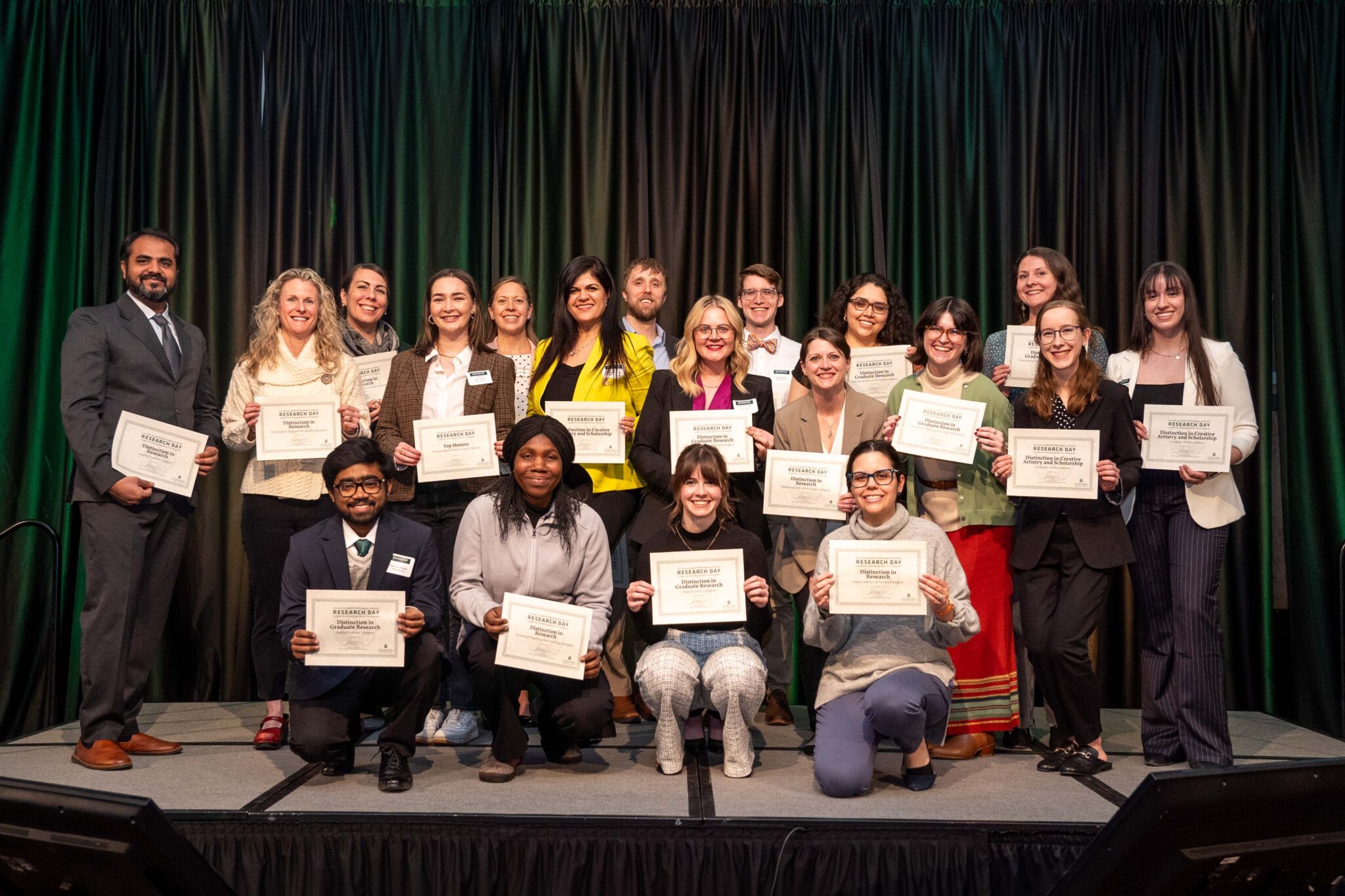 Winners from the Research Day poster presentations stand on stage with their certificates. There are 19 winners.