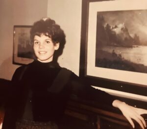 1960s photo of a young woman with short brown hair in a black shirt standing in front of a framed landscape painting