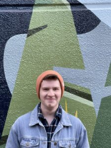 Jack smiles in front of a patterned wall. He is wearing a denim jacket and orange beanie.