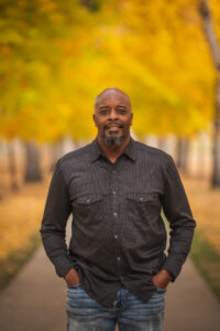 Ray Jackson smiles in his headshot. He is standing in front of orange and yellow fall foliage. He is wearing jeans and gray button up shirt.