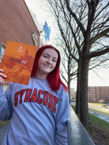 Knittel smiles with her Syracuse acceptance letter. She is wearing a gray sweatshirt that says "SYRACUSE".