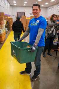 Joel volunteering at the Food Bank of the Rockies. He is wearing a blue shirt and jeans. He is holding a large green bin.