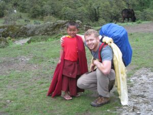 Moon, wearing a backpack, smiles with his arm around a Nepalese child wearing red clothing.