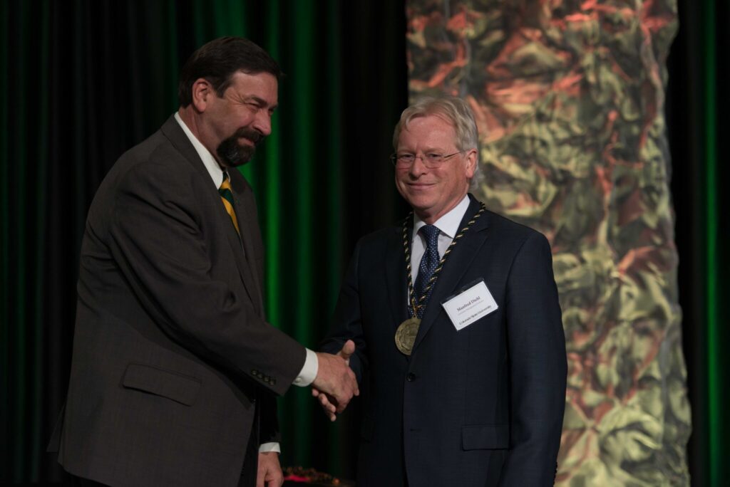 Two men in suits shake hands in front of a decorative background. One is wearing a medal.