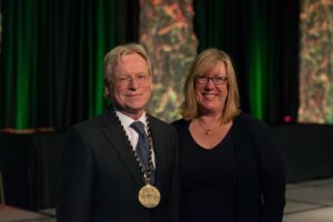 A man in a suit wearing a medal stands next to a woman in front of a decorative background.