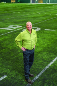 A man in a bright green shirt standing on a football field with the 20-yard-line behind him.