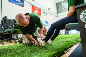 Man in a green shirt on his knees in a lab adjusting sensors on another person's shoes.