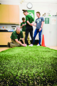 A man kneels to adjust equipment on an athlete's shoe in a lab with green turf on the floor.