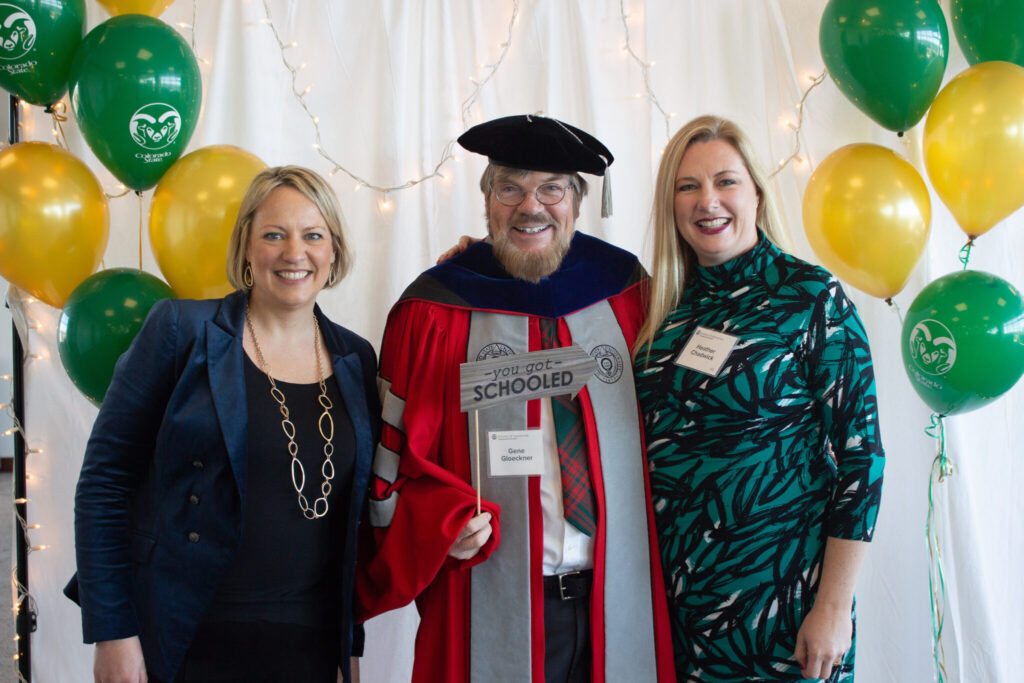 Gene Gloeckner, wearing his academic regalia, smiles with two people at a graduation event, with greena dn gold baloons in teh background.