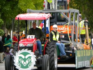 A tractor with a C S U logo on the front pulls a parade float that is construction themed
