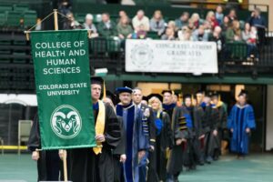 Scott Glick holding a banner that reads "College of Health and Human Sciences, Colorado State University" as he leads a long faculty procession on commencement day. All wearing regalia