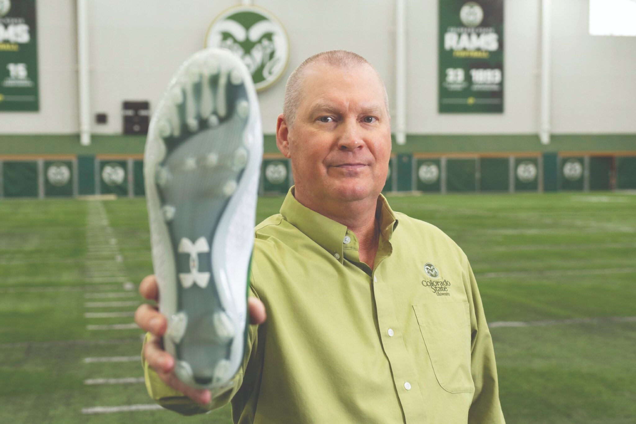 Man in a bright green shirt on an indoor football field holds an athletic shoe with cleats