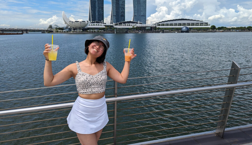 Woman holding two colored drinks against a background with water and buildings