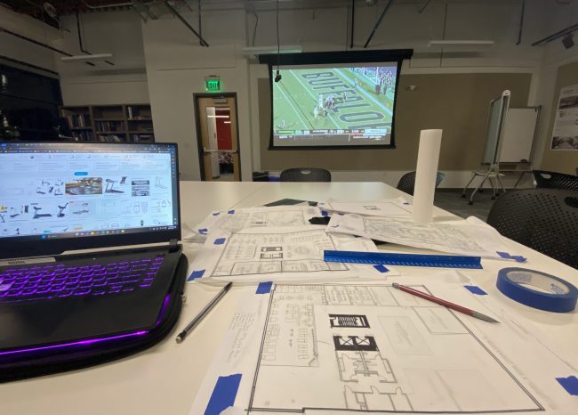 Architectural plans are laid across a desk as a football game plays in the background.