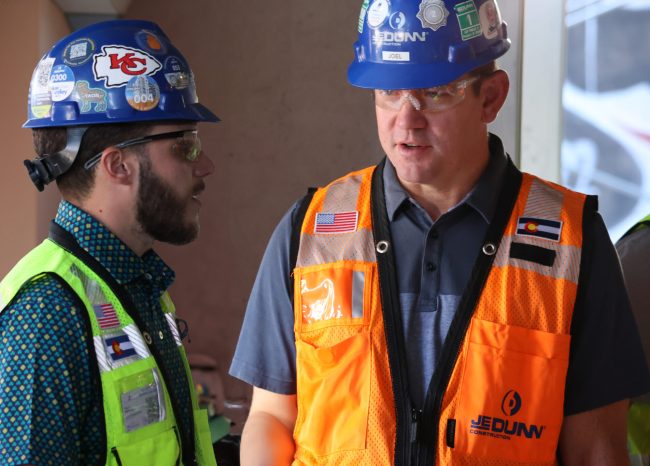 Joel speaks to another man at a construction site. Both are wearing bright safety vests and hard hats.